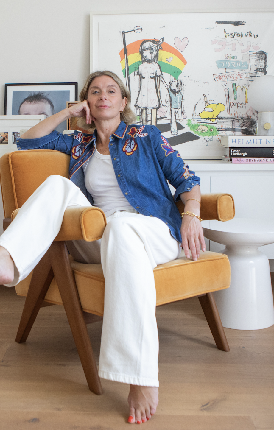 A woman sitting in a mustard-colored armchair in a living room, wearing a blue embroidered denim shirt and white pants, with her right hand supporting her head and her left resting on the armrest. Behind her is a whiteboard with a child's drawing of a person with a rainbow, and books on a white sideboard.