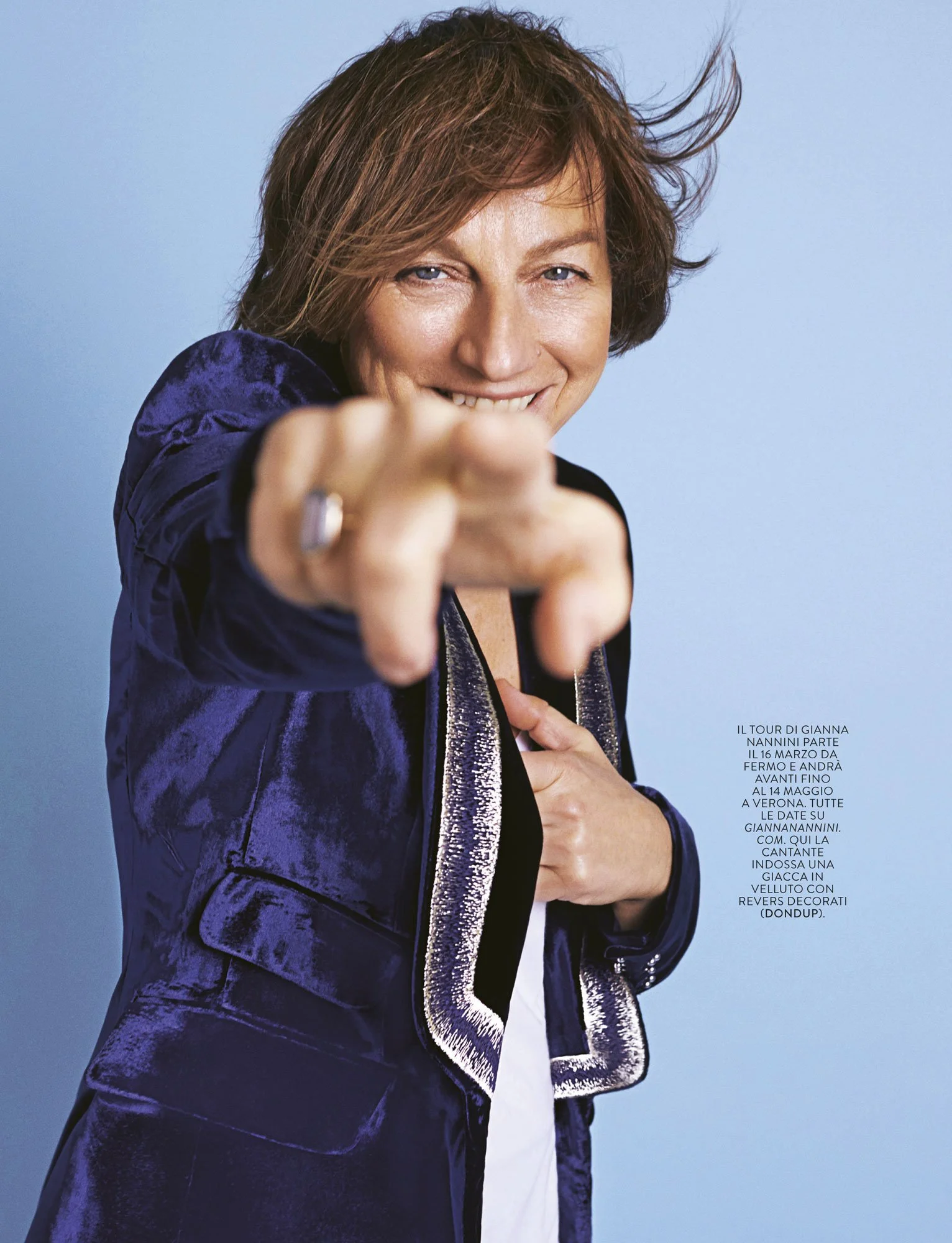 A woman with short, tousled brown hair pointing directly at the camera, smiling, dressed in a navy velvet jacket with silver details, standing against a light blue background.