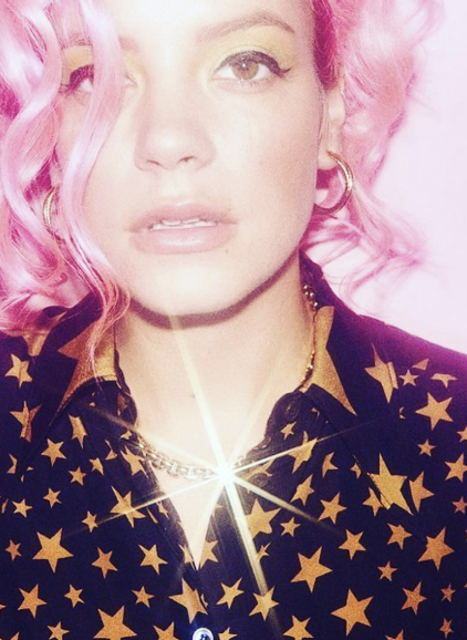 A woman with pink curly hair, one eye closed, wearing gold hoop earrings and a star-patterned black shirt, with a star-shaped light reflection on her chest.