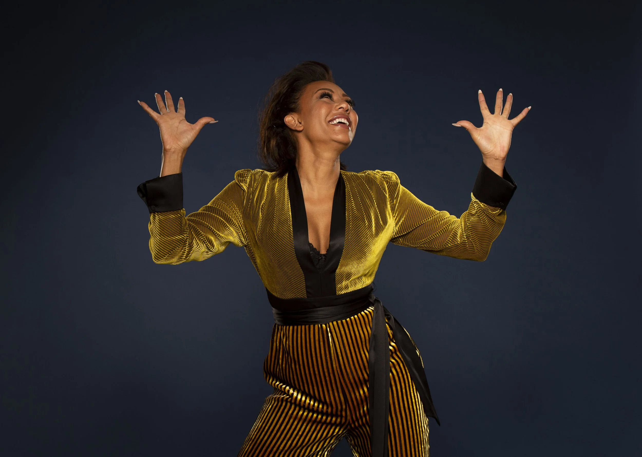 A woman with short dark hair, wearing a shiny yellow and black striped suit, is smiling and laughing with her arms raised against a dark background.