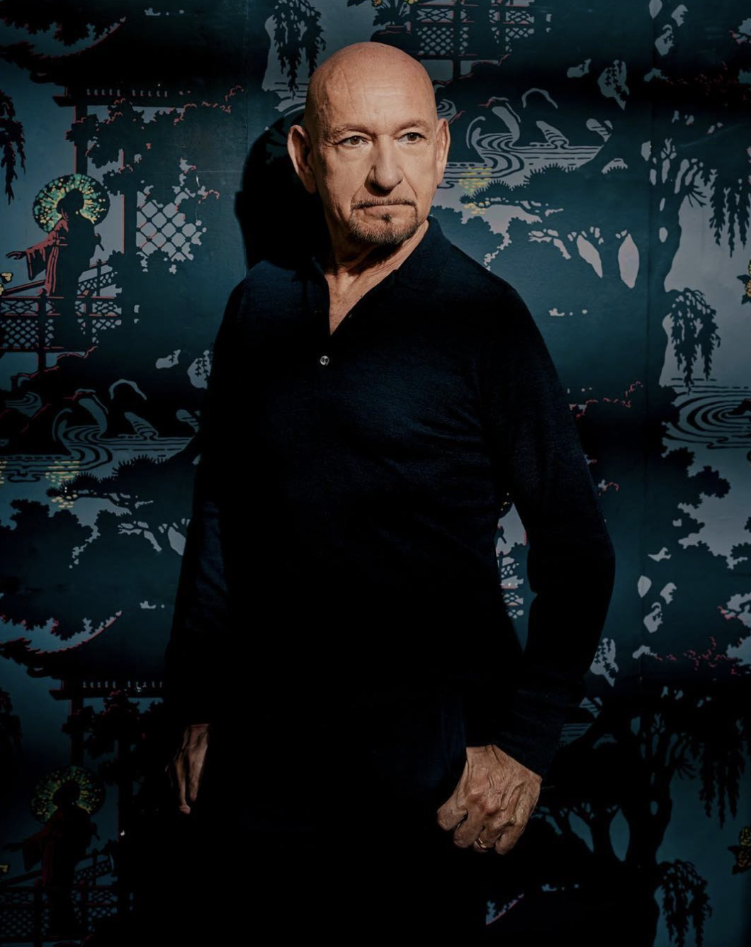 A man with a shaved head and goatee wearing a black shirt stands in front of a blue and black Asian-themed mural.