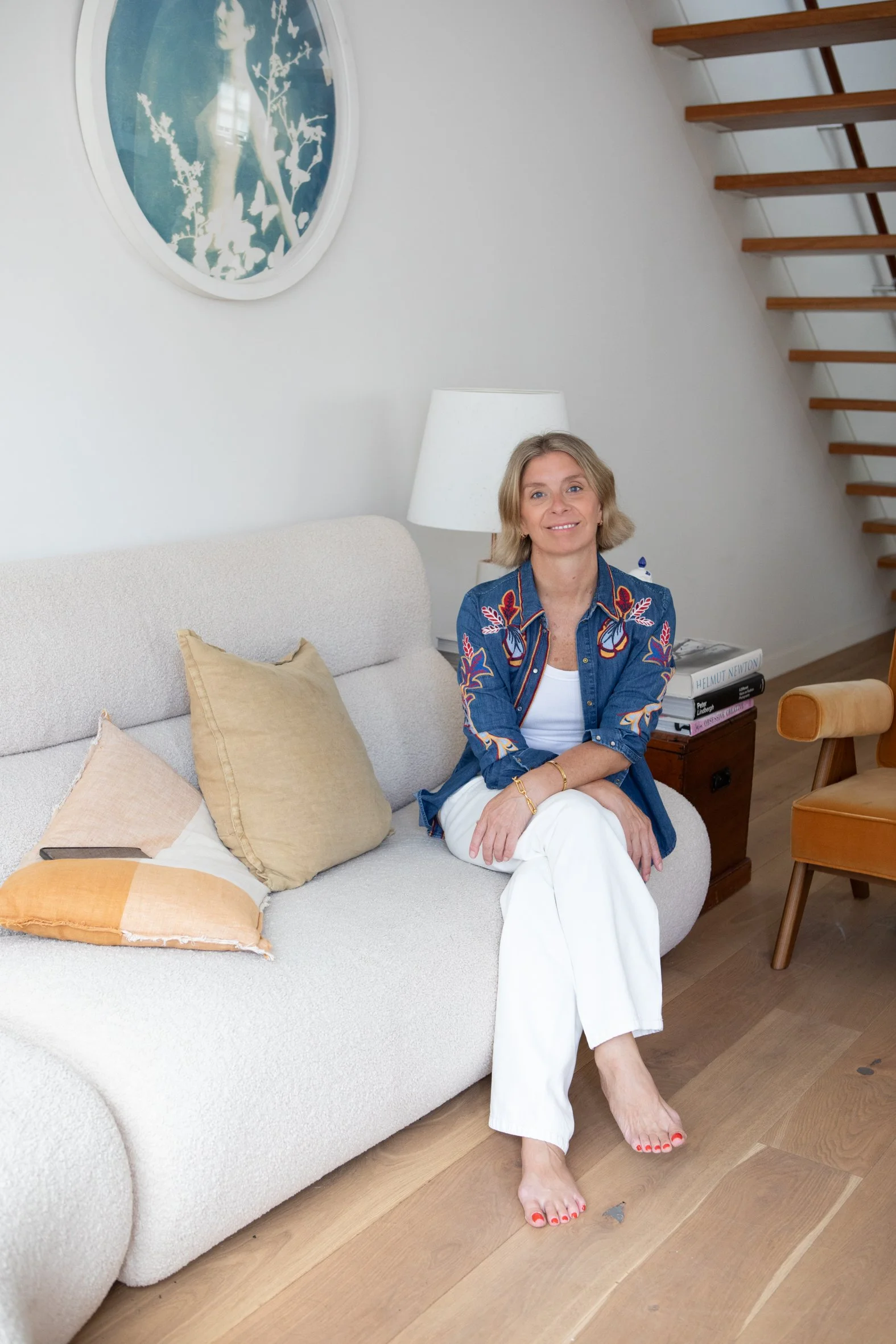 A woman sitting on a cream-colored couch in a living room, smiling, wearing a blue embroidered jacket and white pants.