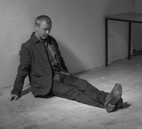 Man sitting on the floor against a wall in a room with a desk nearby