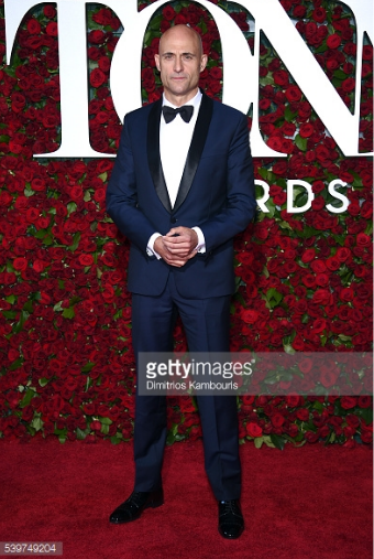 Man in a navy blue tuxedo with black lapels, white shirt, and black bow tie standing on a red carpet in front of a background with red roses and large white letters.