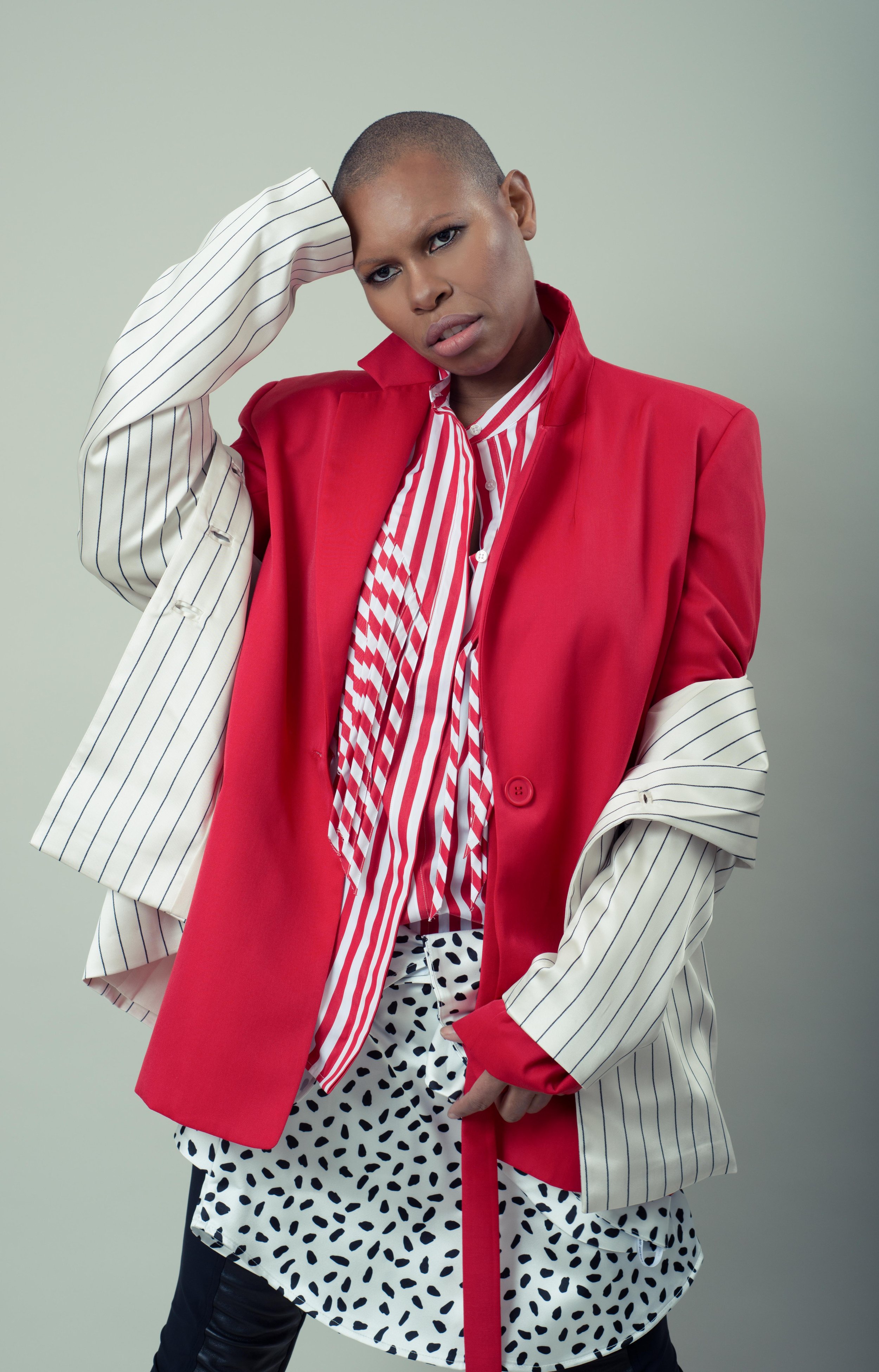 A person with short hair wearing a red blazer over a striped shirt and pants with black spots, standing against a plain background.