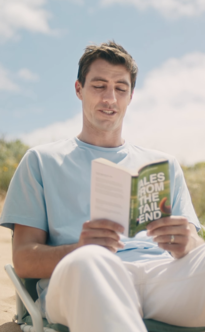 A man reading a book outdoors on a sunny day with a partly cloudy sky, sitting on a beach chair, wearing a light blue shirt and white pants.