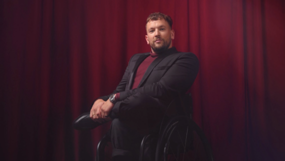 Man in a wheelchair wearing a black blazer and maroon shirt, sitting in front of red curtains.
