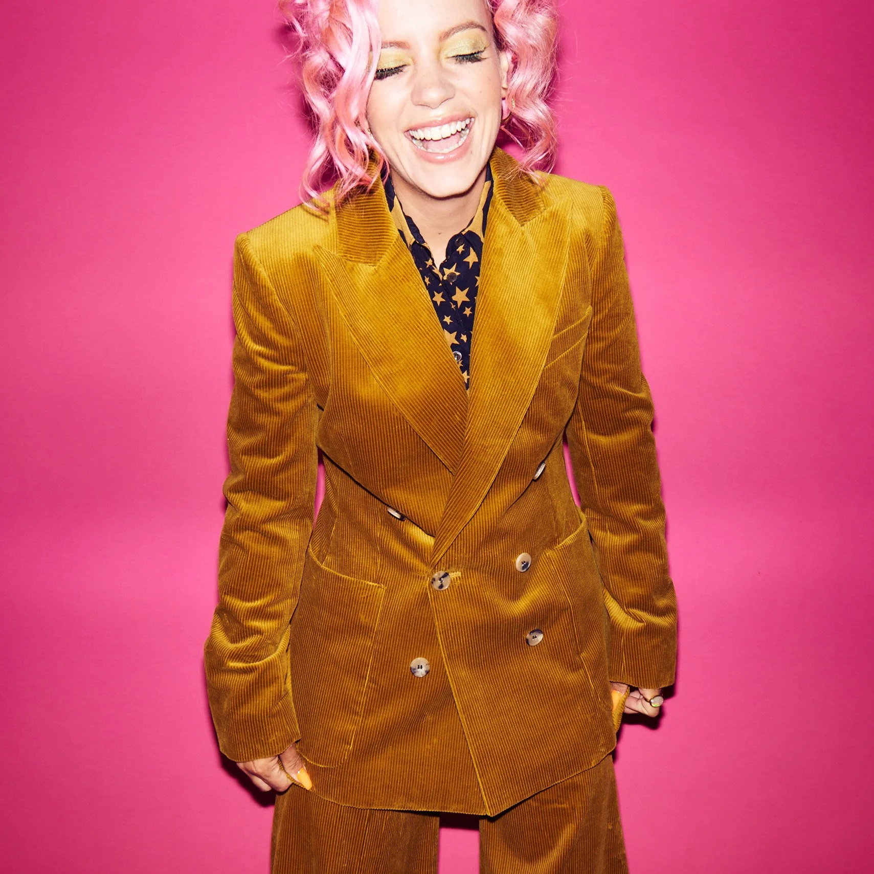 A woman with curly pink hair, yellow eyeshadow, and a wide smile, wearing a yellow corduroy blazer and matching pants, standing against a pink background.