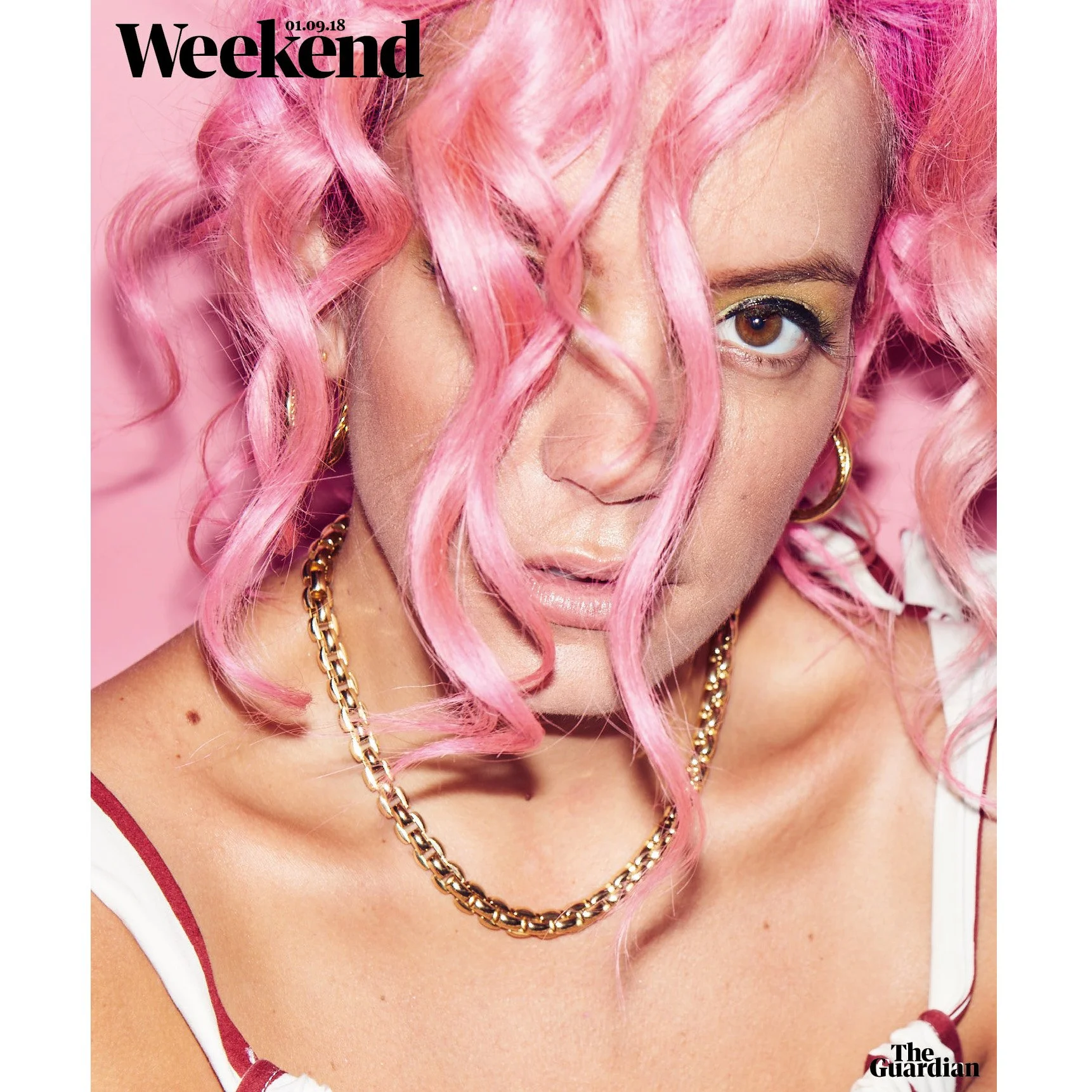 Close-up of a woman with curly pink hair covering parts of her face, wearing gold jewelry, and a white top with red straps, on the cover of Weekend magazine, dated September 1, 2018.