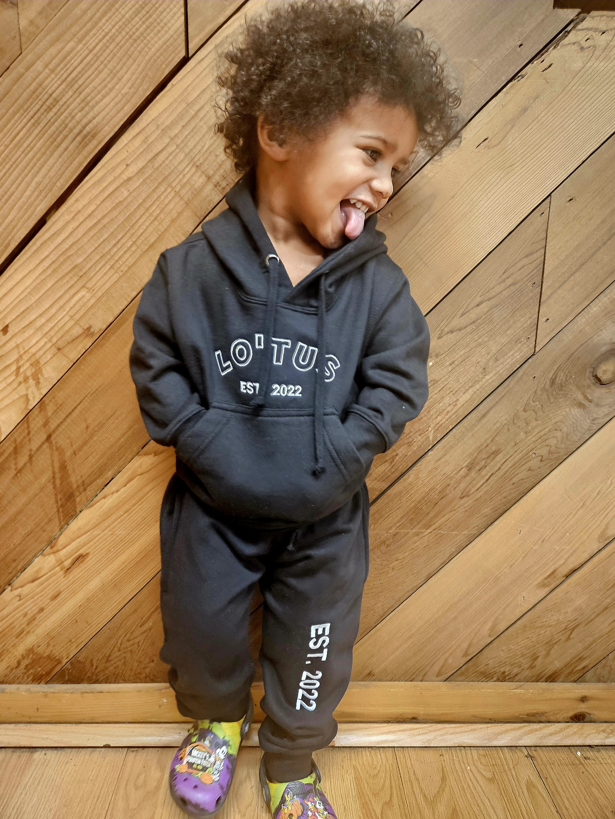 A young boy with curly hair sticking out his tongue, wearing a black hoodie and matching sweatpants with 'L.O.T.U.S EST 2022' printed on them, standing against a wooden wall.
