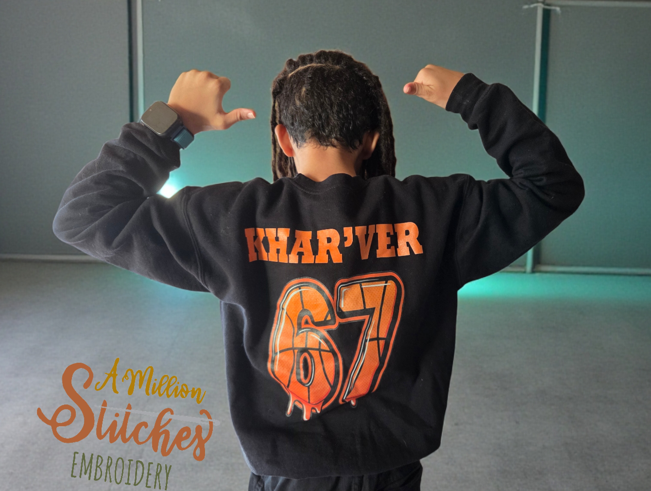 Back of a child wearing a black hoodie with 'KHAR'VER' and '67' printed on it, flexing arms with fists near head, inside a room with teal walls.