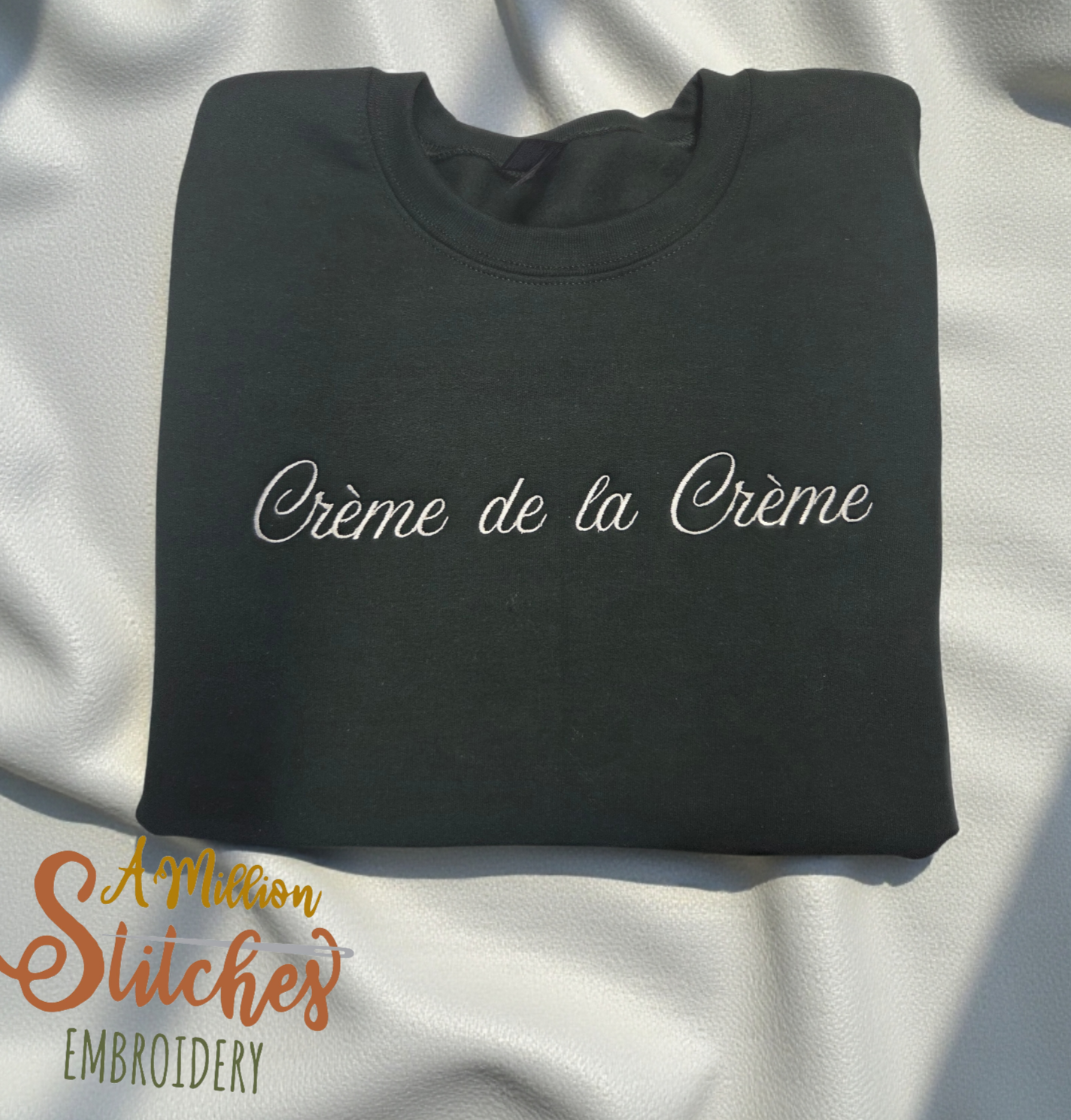 forest green sweater with white embroidered cursive text 'Crème de la Crème' on the front, placed on a white textured surface.