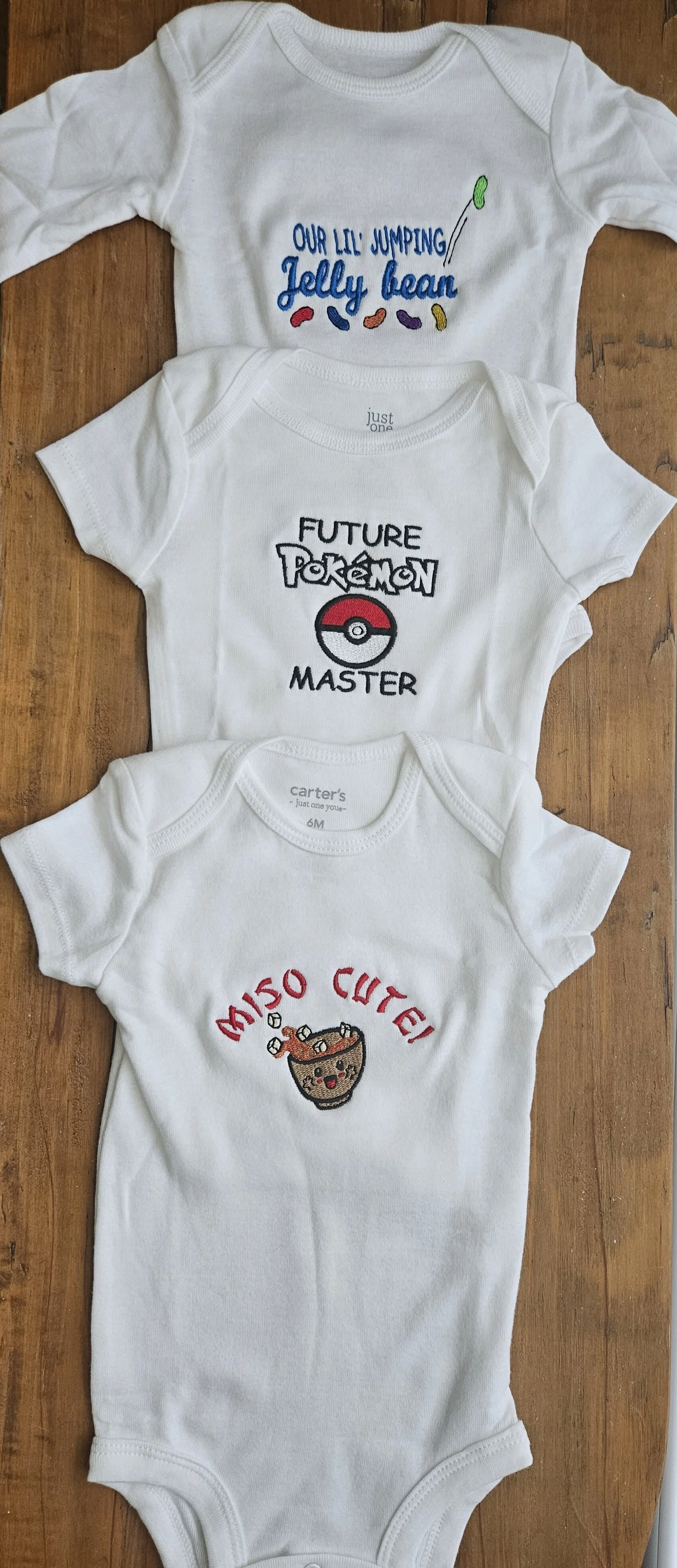 Three white baby onesies laid flat on a wooden surface, each with embroidered or printed designs and text.