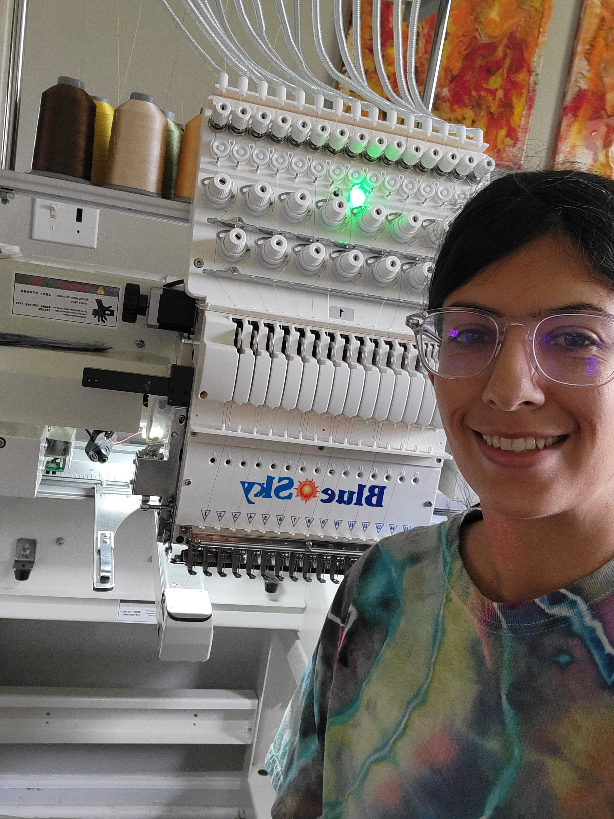 A smiling woman with glasses takes a selfie in front of an embroidery machine with spools of thread and a green light on.