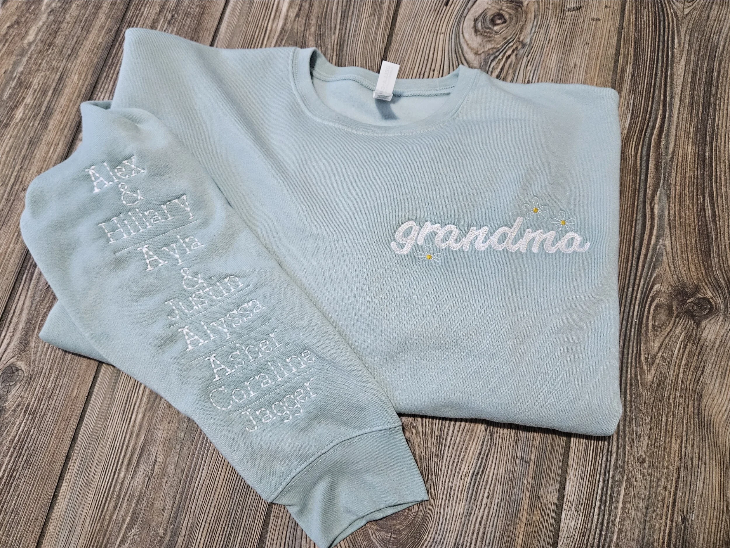 A light blue sweatshirt with the word 'grandma' embroidered on it, and a light blue sleeve with embroidery of the names of grandchildren, Alex, Ethan, Anya, and others, laid flat on a wooden surface.