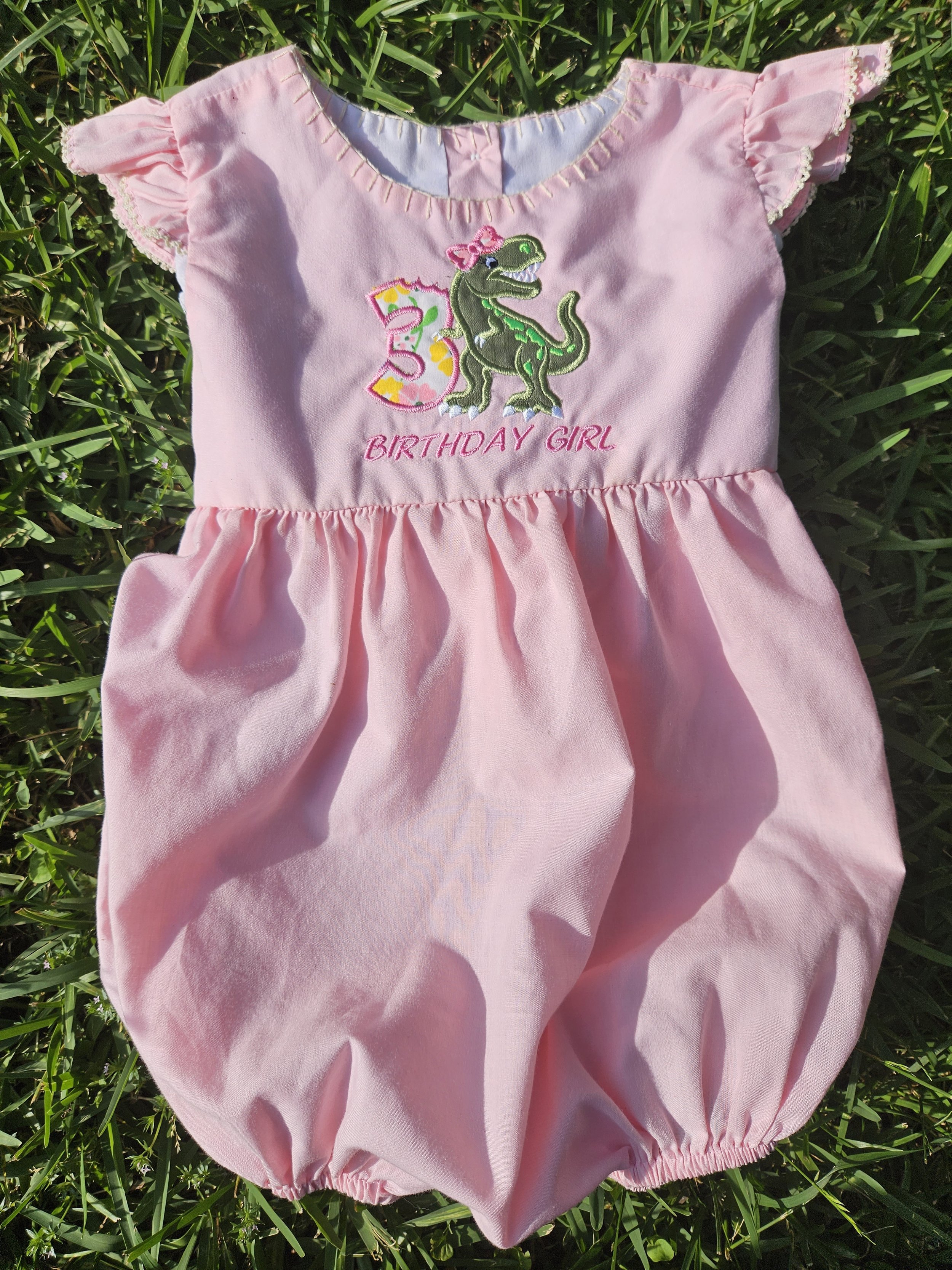Pink baby girl's birthday dress with embroidered dinosaur and '3' and 'BIRTHDAY GIRL' text, lying on grass.
