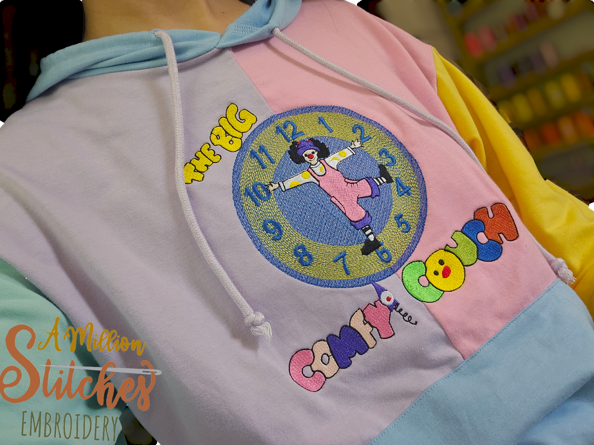 Close-up of a colorful hoodie with a clock, cartoon girl, and the words "Time & Big" and "Complete" embroidered on it. The hoodie has pastel colors, including pink, yellow, blue, and purple. the big comfy couch