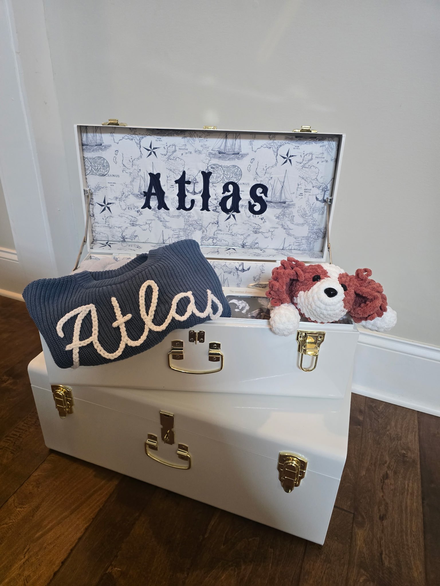 A white trunk with gold hardware and a chest on top, containing a navy blue knit blanket with 'Atlas' in white letters and a plush dog toy with reddish-brown floppy ears and white face, in a room with wooden floor and white wall.