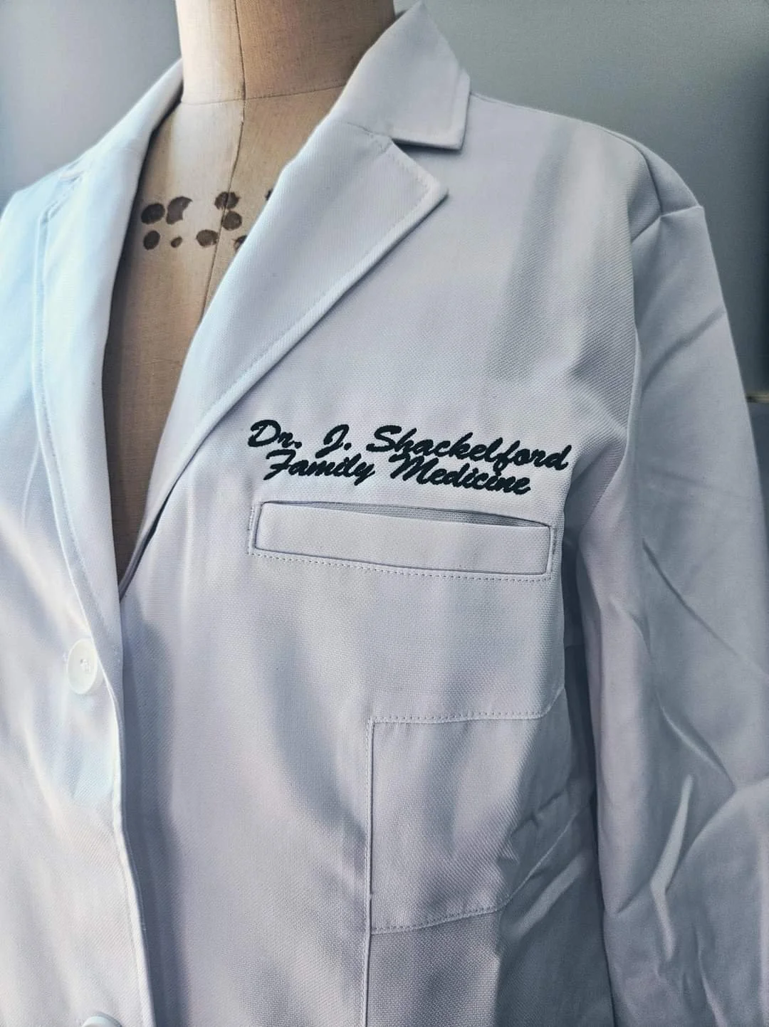 White lab coat with embroidered text 'Dr. J. Shackelford, Family Medicine' on the chest pocket, displayed on a mannequin.