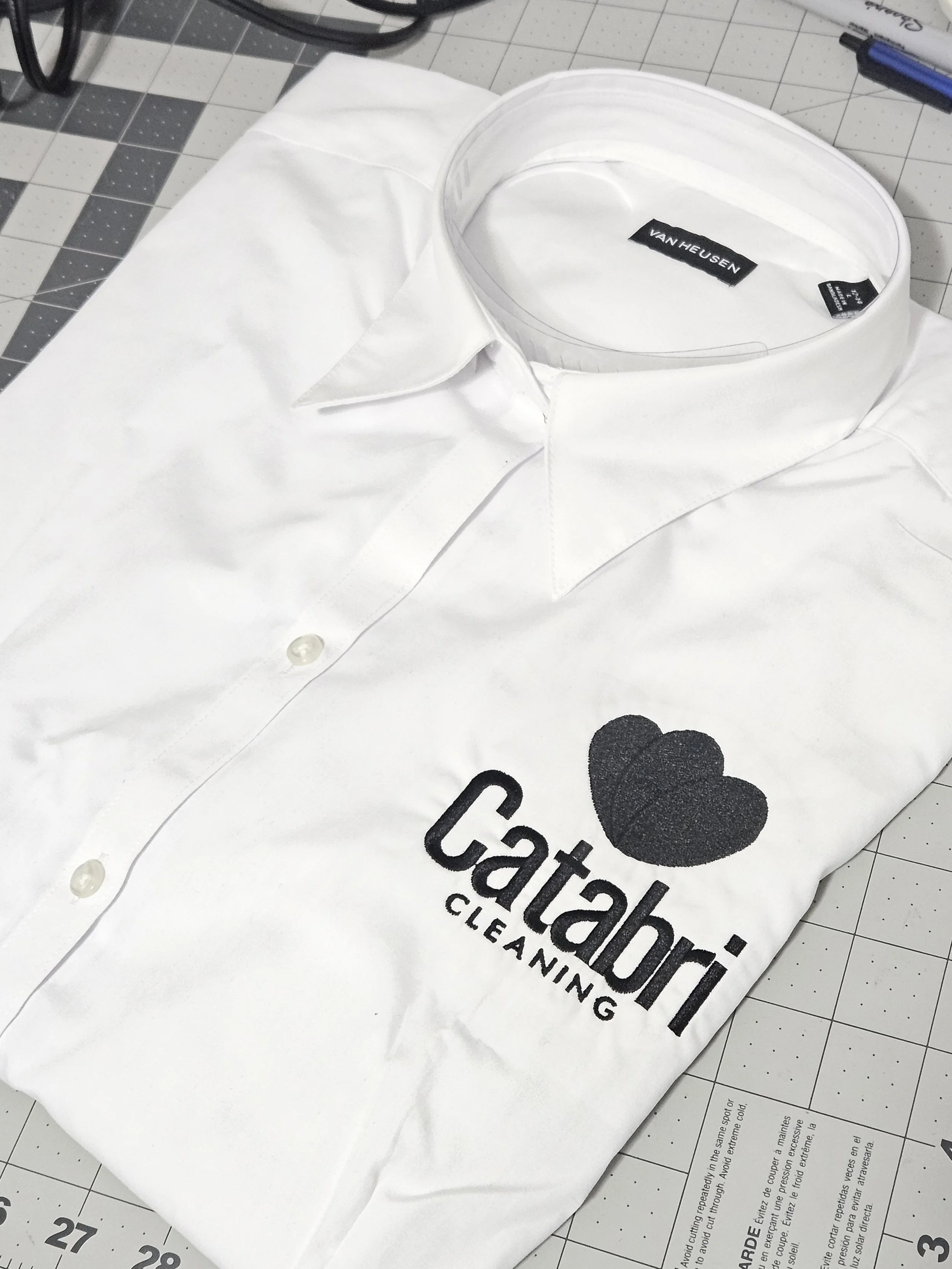 White dress shirt with a logo that says "Catabri Cleaning" and has a black heart symbol embroidered on the chest.