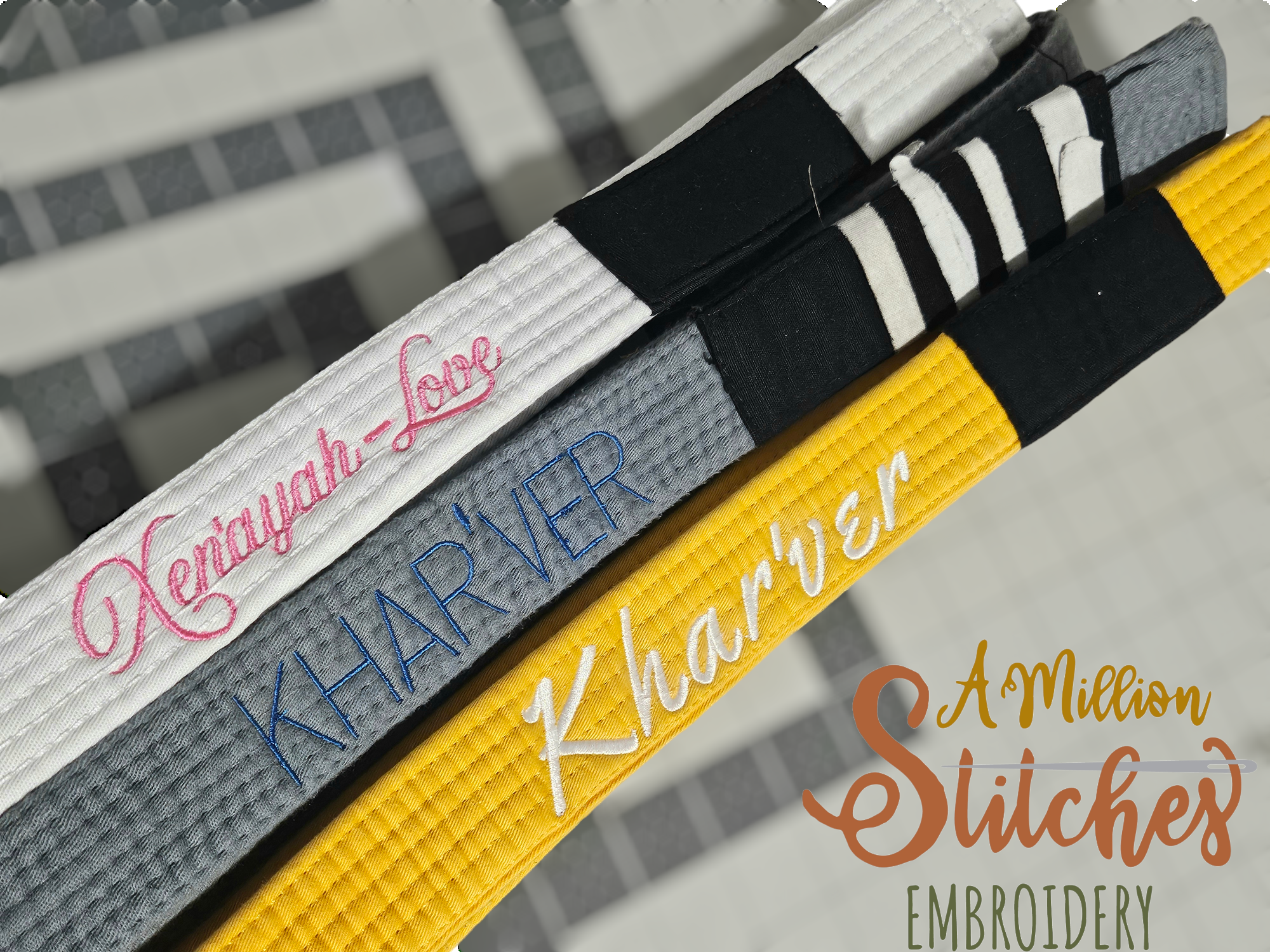 Close-up of embroidered jiu jitsu belts with names 'xen'ayah-Love,' 'KHARVER,' and 'Khar'Ver,' featuring decorative stitching in pink, blue, and white, arranged on a black and yellow surface.