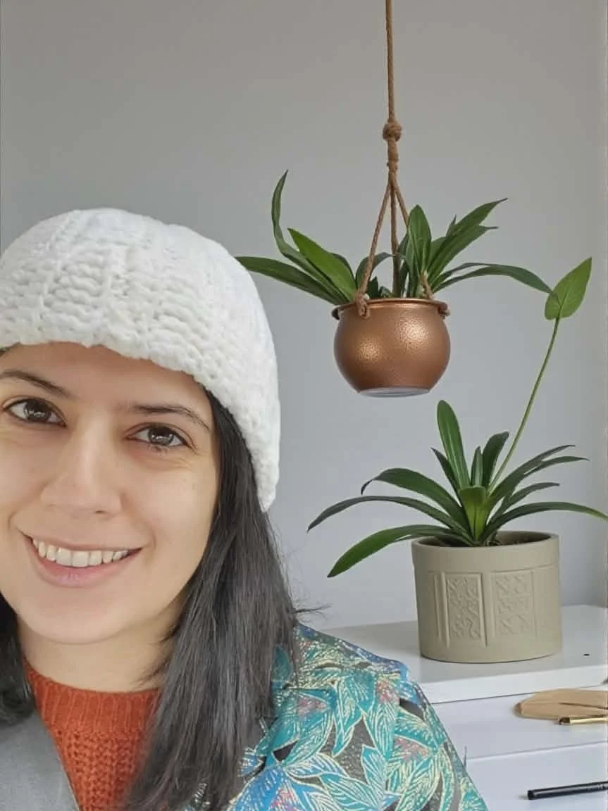 A woman smiling, wearing a white knit hat and an orange sweater, standing next to two potted plants on a white table.