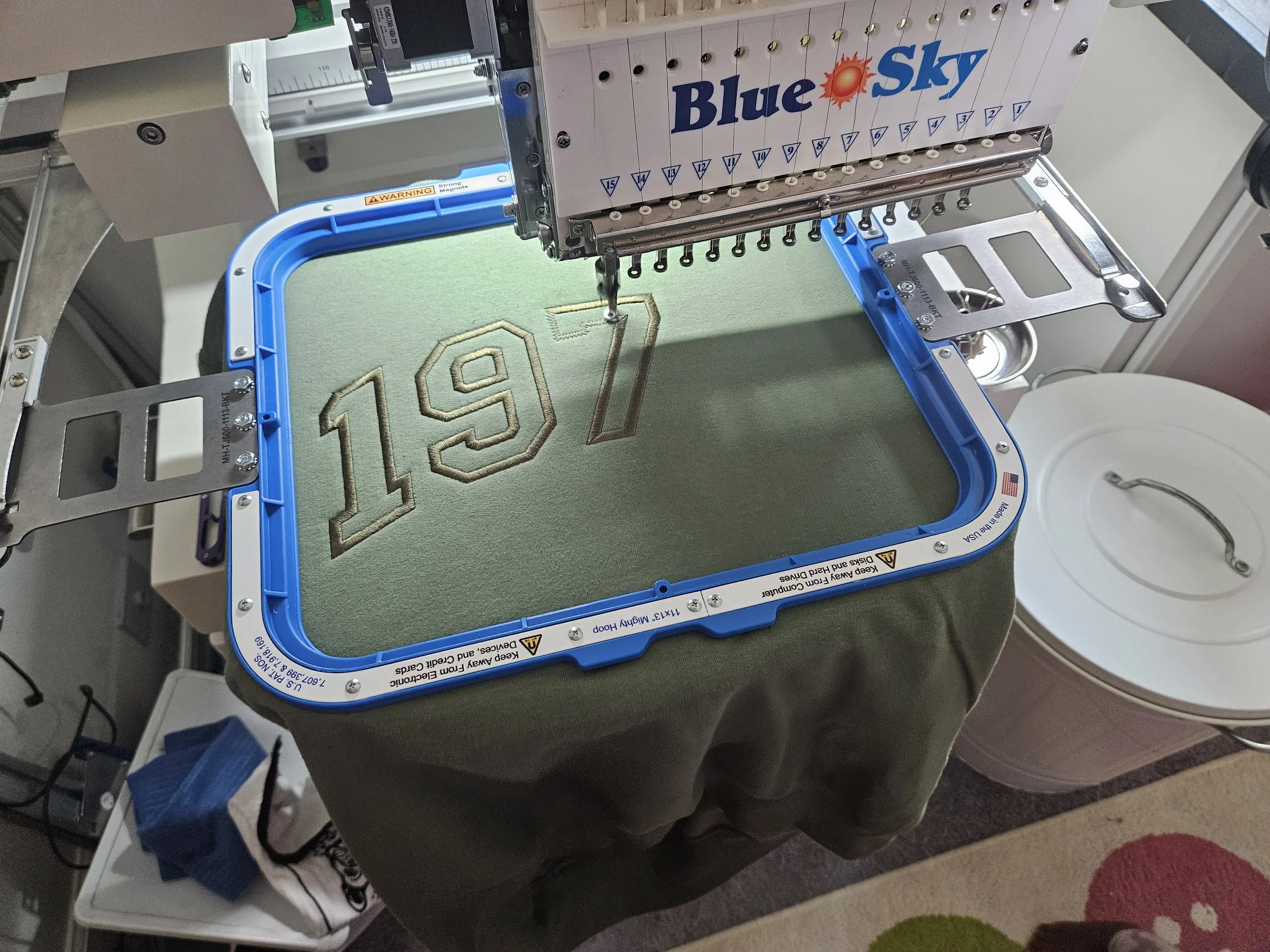 Embroidery machine stitching the numbers '197' onto a green fabric surface.