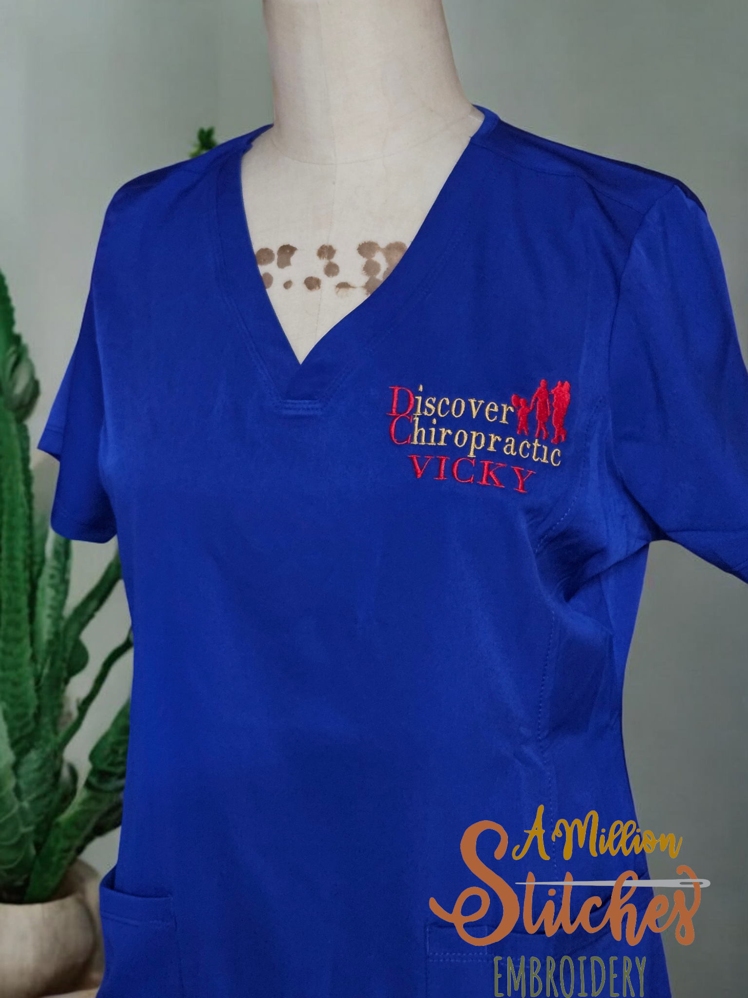 Blue scrub top with embroidered logo and name, displayed on a mannequin with a plant in the background.