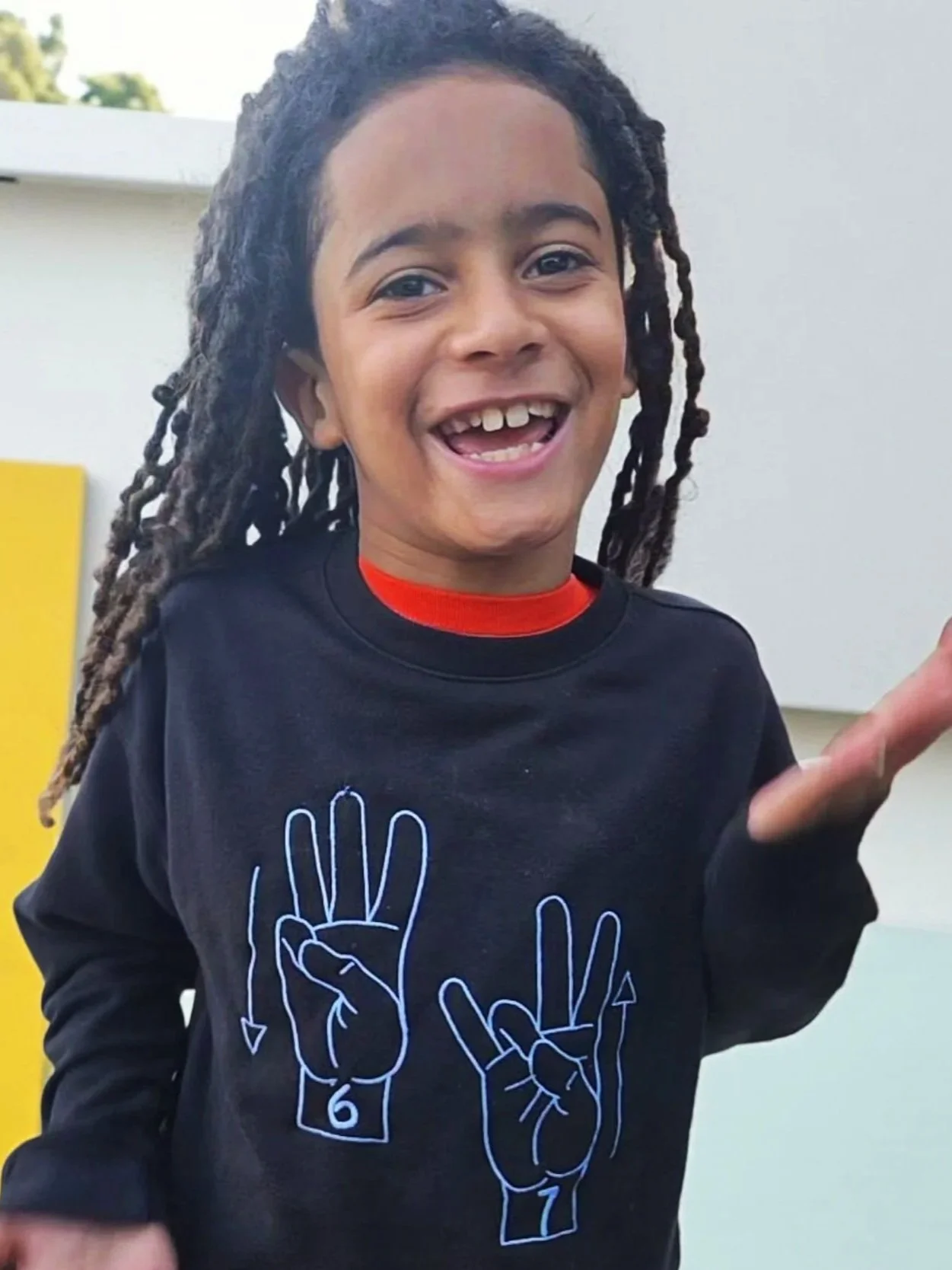 A young boy with long locs smiling and waving outdoors, wearing a black sweatshirt with white embroidered 6,7 in sign language