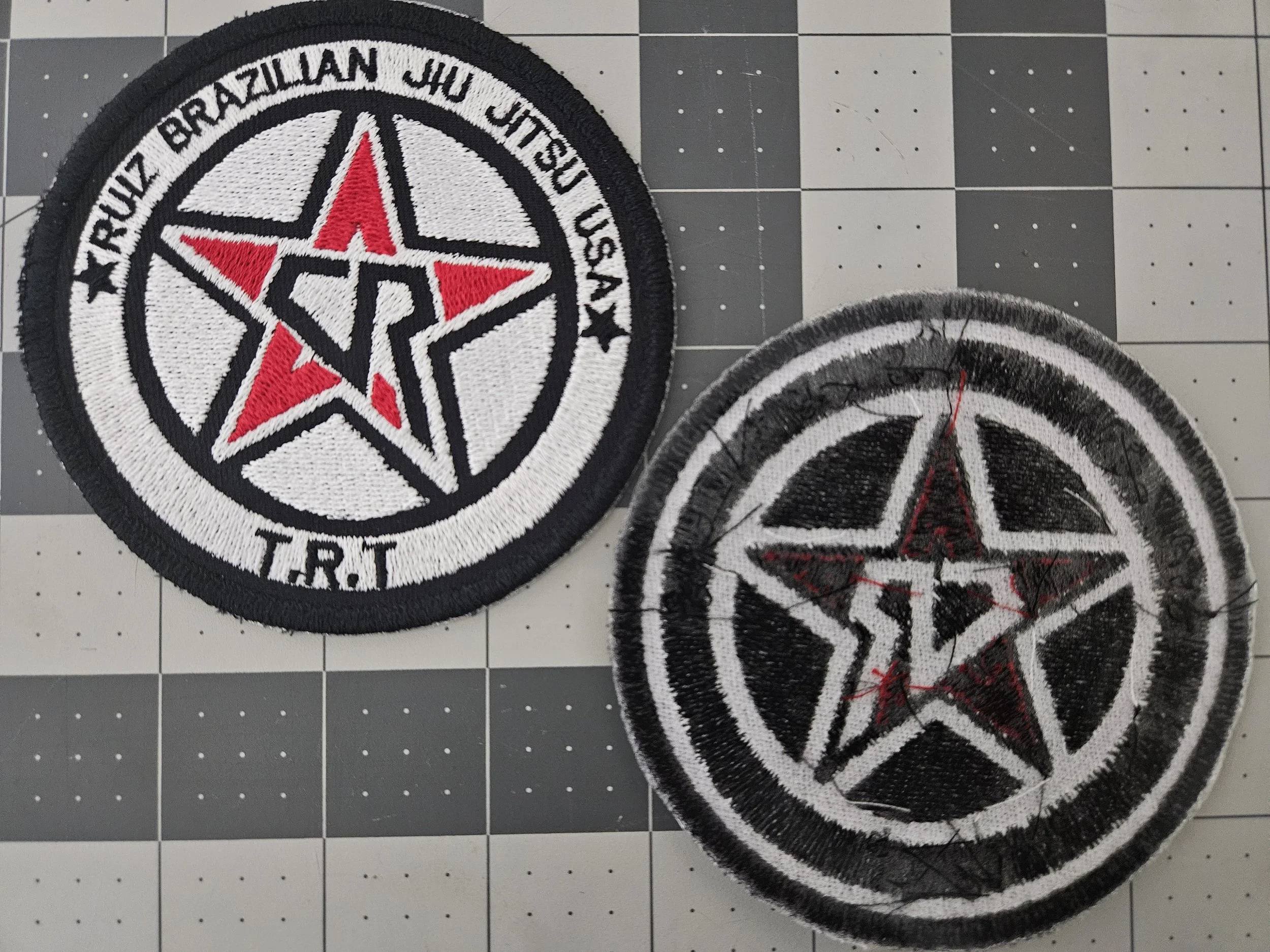 Two embroidered patches with the emblem of the True Brazilian Jiu Jitsu U.S.A., featuring a red and black star with a pentagon inside, surrounded by a white circle with black borders. One patch is clean, the other is worn and frayed.