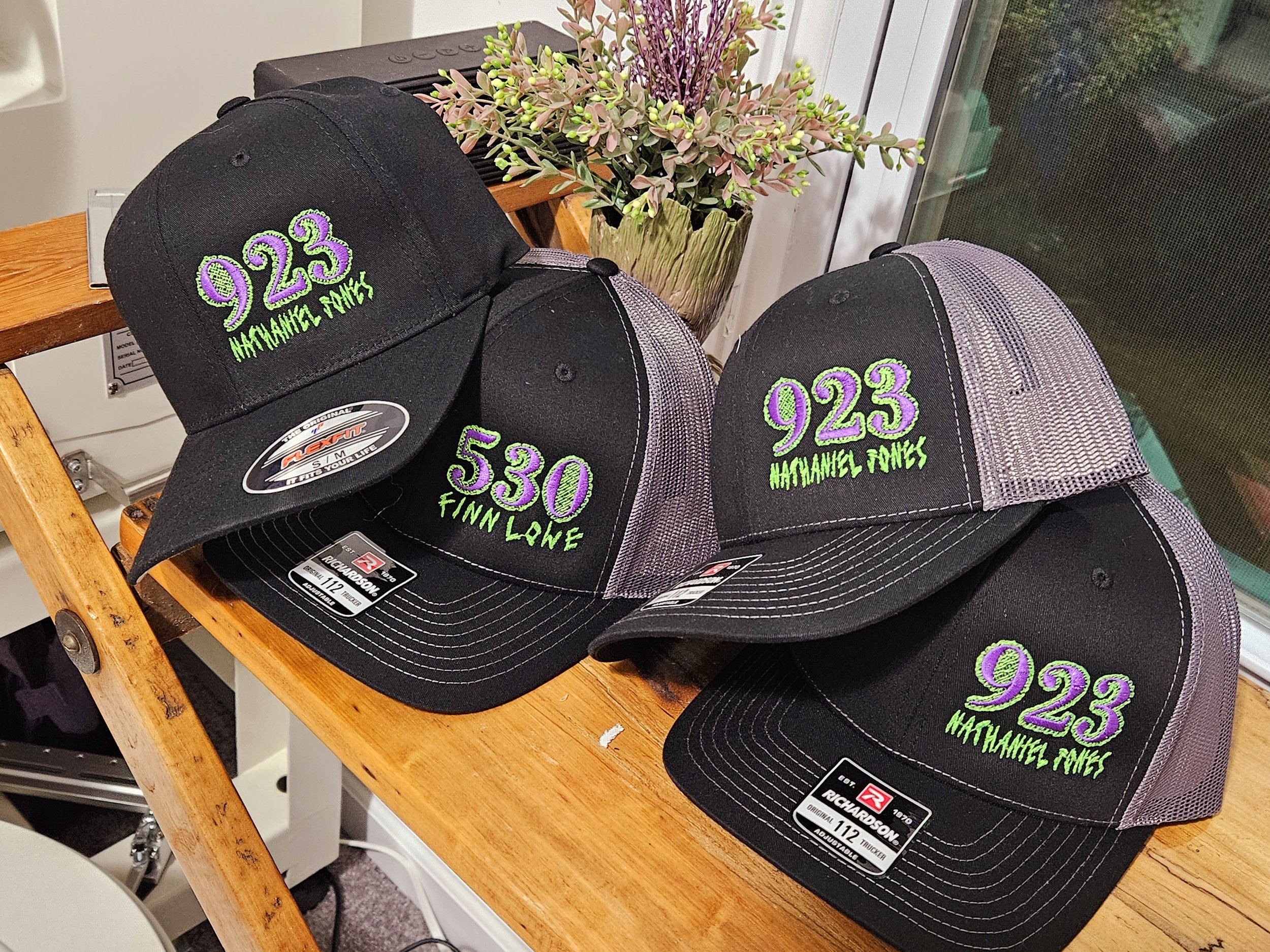 Three black and gray trucker hats on a wooden shelf, each embroidered with the number '923' and the name 'HATCHANEL FONES' in green; one had a sticker that says 'RICHARDSON 112.'