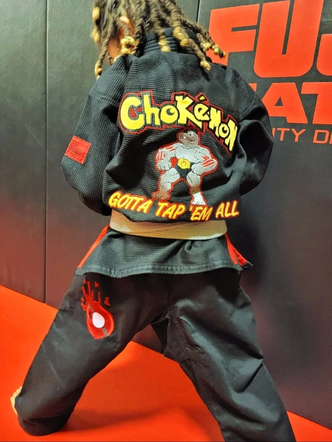 Child in martial arts uniform with a black gi, standing on a red mat, wearing a black jacket with colorful embroidery of a fighting pokemon and the text 'Choke' and 'Gotta Tap 'Em All', with a background featuring the logo of a martial arts gym.