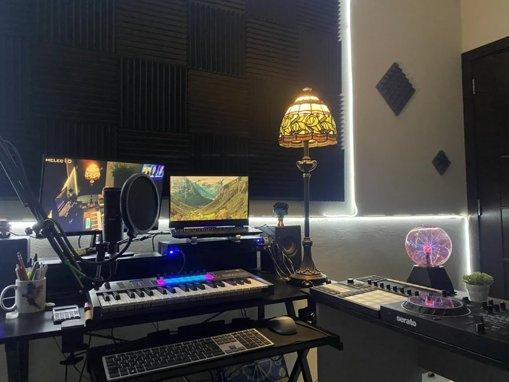 Music studio with two computer monitors, a keyboard, a mixing console, a microphone, a vintage-style lamp, a plasma ball, and soundproof foam panels on the walls.