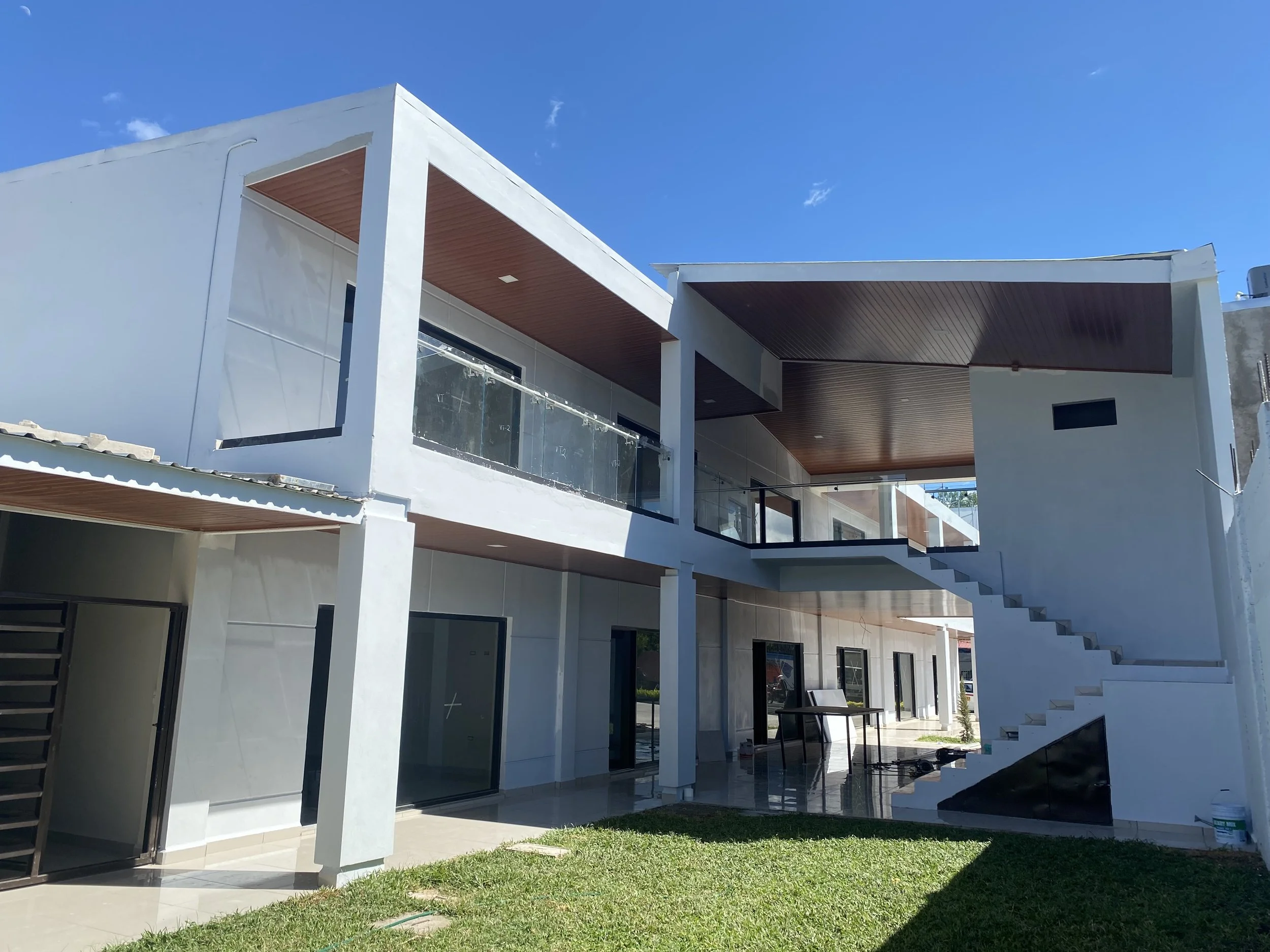 Modern white building with multiple floors, large windows, and a balcony with glass railing, set against a bright blue sky.