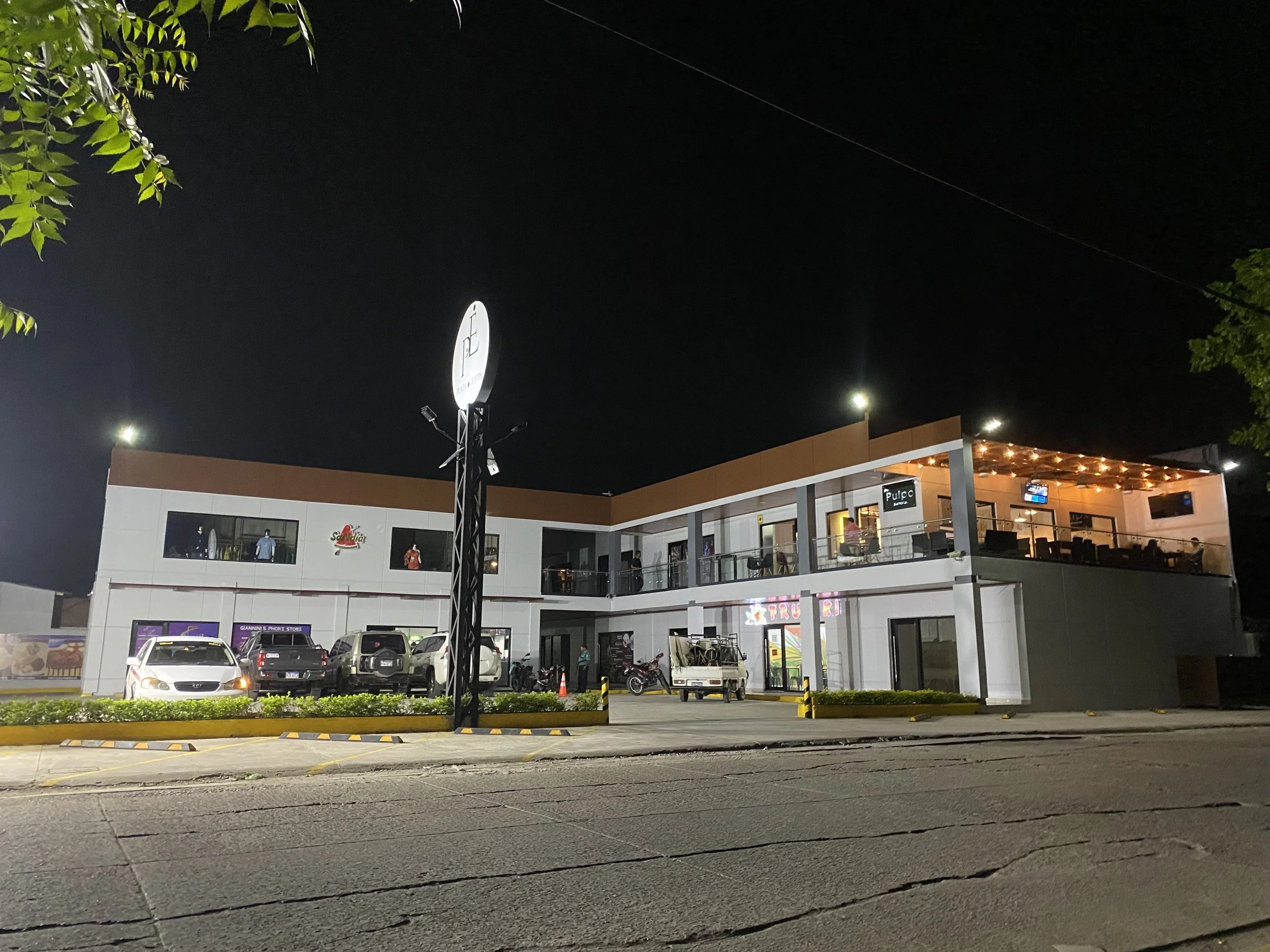 Night view of a commercial building with a parking lot, several parked cars and motorcycles, trees, and streetlights illuminating the area.