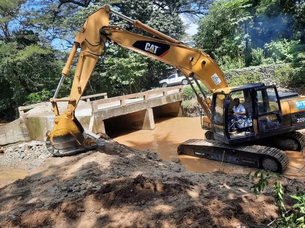 A construction excavator working in a muddy river, with a small bridge and green trees in the background.