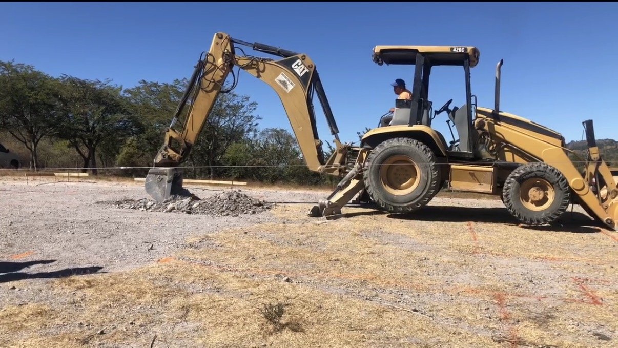 A construction site showing a large yellow Caterpillar excavator with a woman operator, digging into the ground on a clear, sunny day with blue sky and trees in the background.
