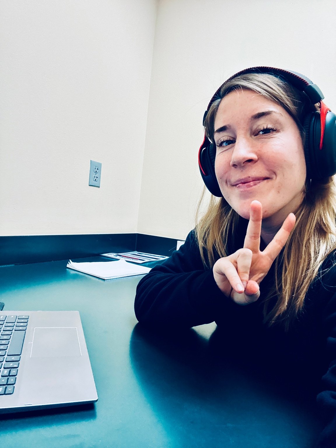 Young woman with long hair smiling and making a peace sign while wearing large headphones, sitting at a desk interpreting with a laptop and papers in a plain office.