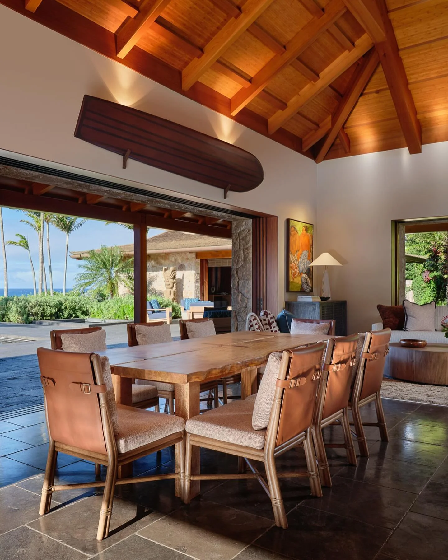 A dining area with a wooden table and eight chairs, open to an outdoor patio with tropical plants, palm trees, and a view of the ocean.