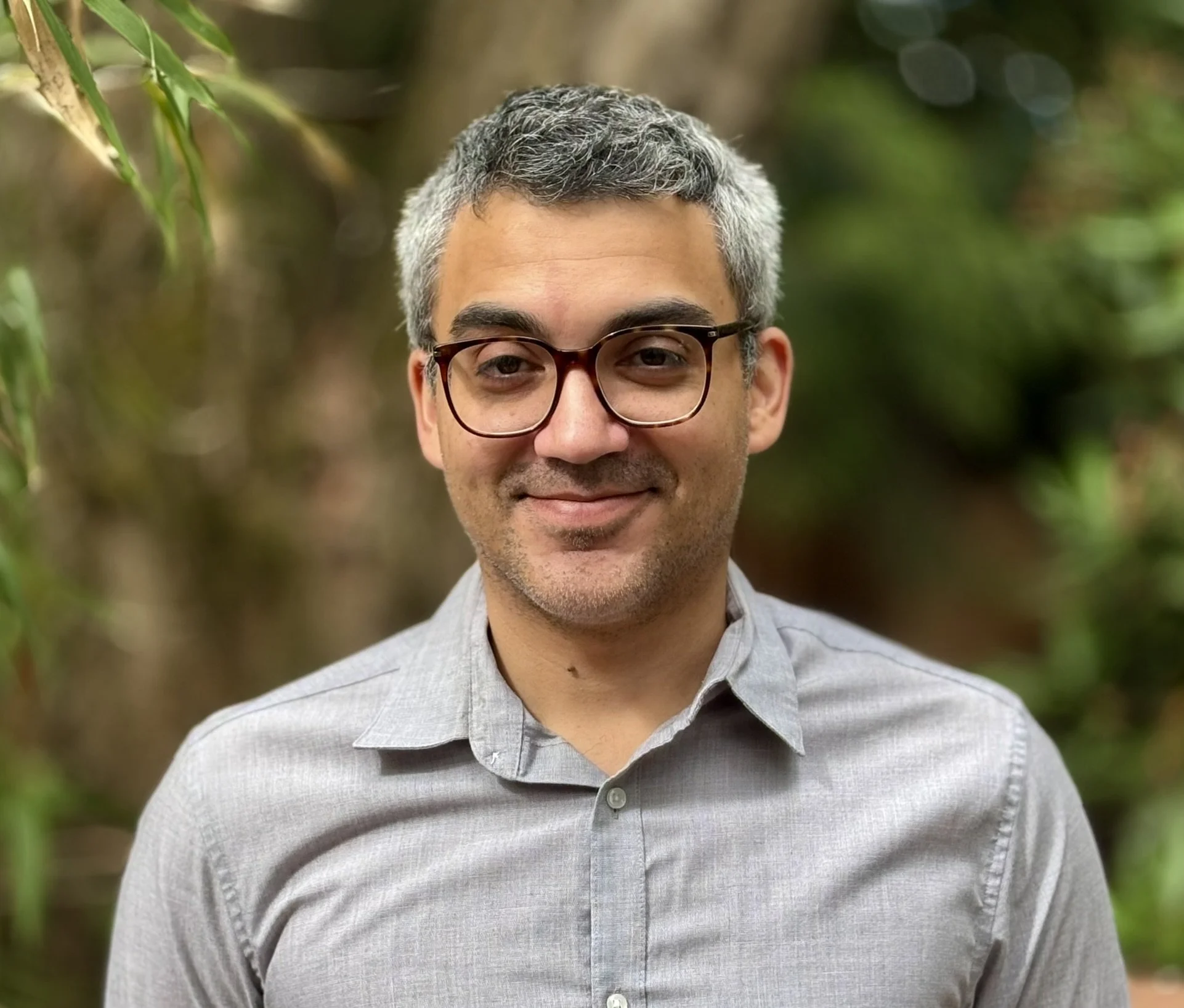 Close-up of a smiling man with gray hair and glasses, wearing a light gray shirt, outdoors among green foliage.