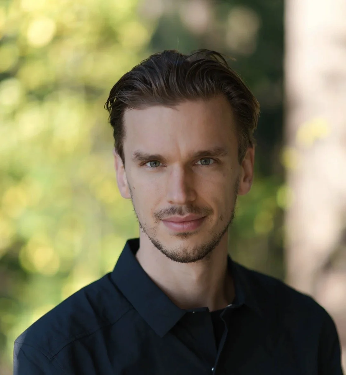 Close-up portrait of a man with brown hair, blue eyes, and light facial hair, wearing a black shirt, outdoors with blurred green and yellow foliage in background.