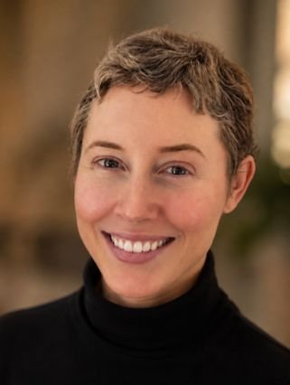 A smiling woman with short curly hair wearing a black turtleneck, indoors with warm lighting.