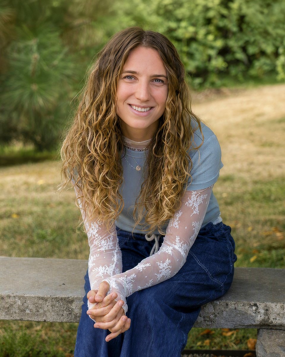 A young woman with long, curly blonde hair, wearing a light blue top with sheer embroidered sleeves, sitting on a concrete bench outdoors with greenery in the background, smiling at the camera.
