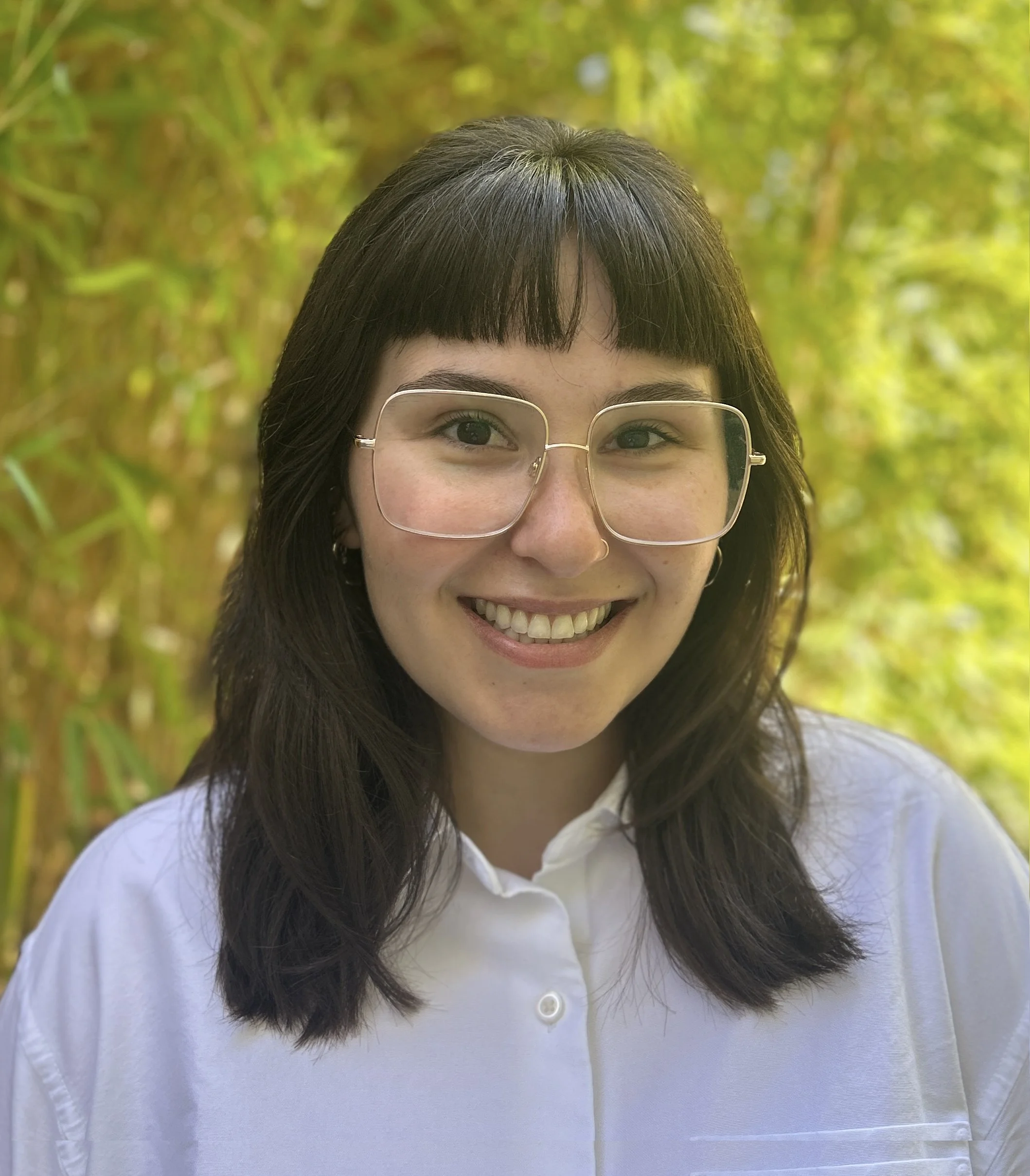 Young woman with dark hair, glasses, and a white collared shirt smiling with green and yellow blurred foliage in the background.