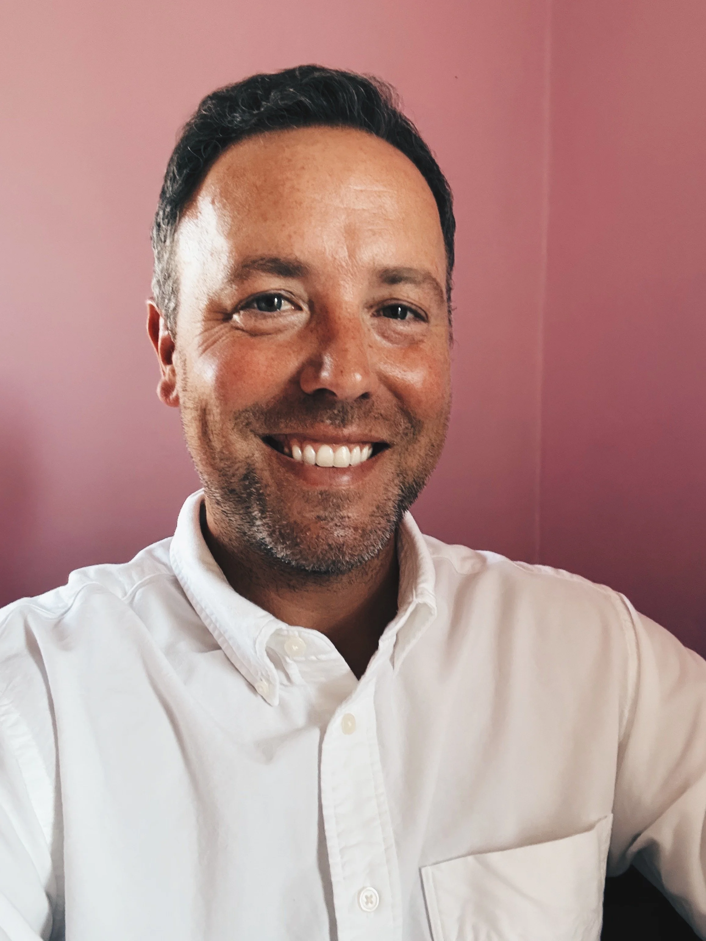 Close-up of a smiling man in a white dress shirt, against a pinkish wall.