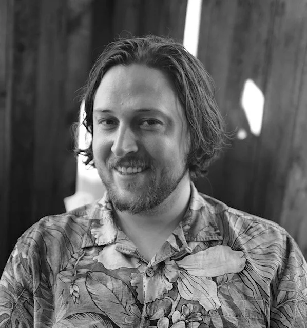 A young man with wavy hair and a beard, smiling, wearing a tropical shirt, in front of a wooden background.