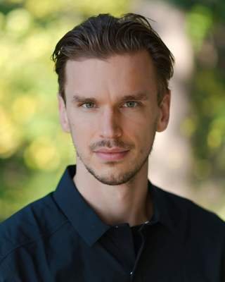 A young man with brown hair and a slight beard, wearing a dark blue shirt, standing outdoors with blurred green foliage in the background.