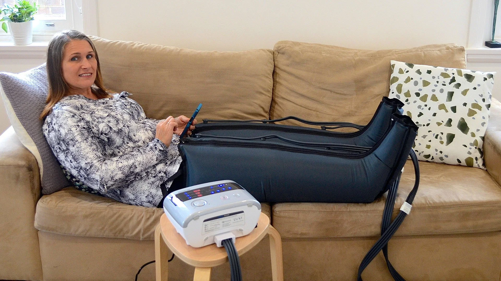 A woman lying on a beige couch with a pillow, using her phone, with a compressed air compression device on her lap and a machine on a small wooden table connected to a large black inflatable device that covers her legs.