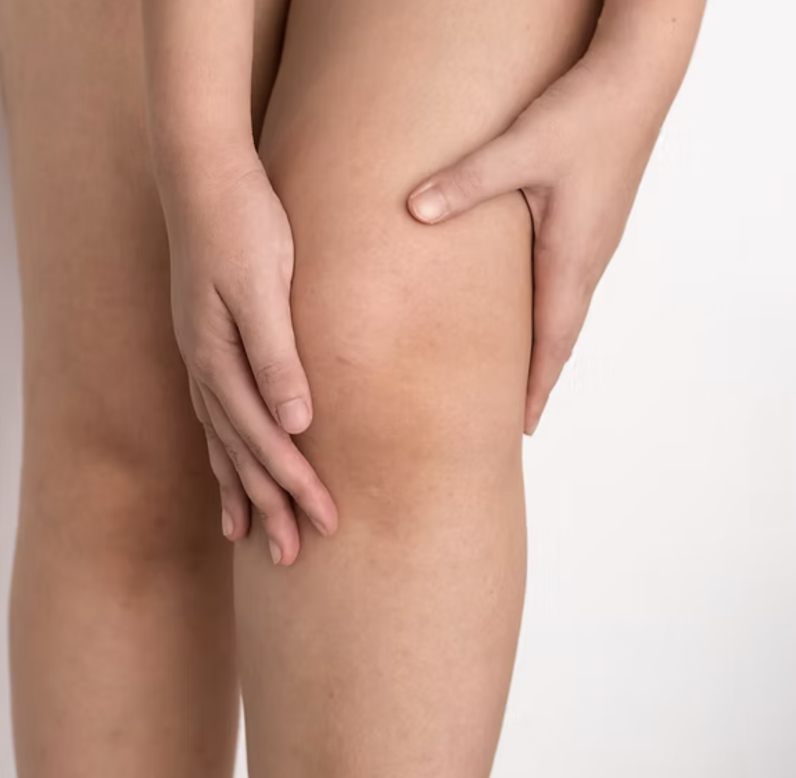Close-up of a person's nude body, focusing on the thigh and hand covering part of the leg with a neutral background.
