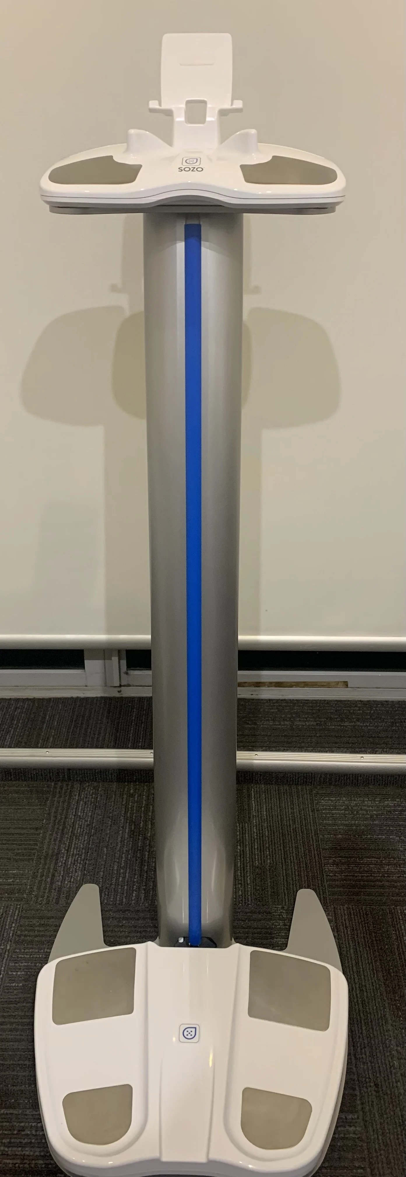 Standing medical scale with a tall, vertical support, digital display, and platforms for weighing, in a room with carpeted floor and plain wall.